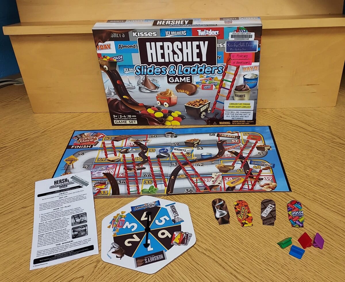 A Hershey Slides & Ladders board game set on a wooden table, showing the game box, game board, spinner, instruction sheet, colorful character tokens, and four player pieces.