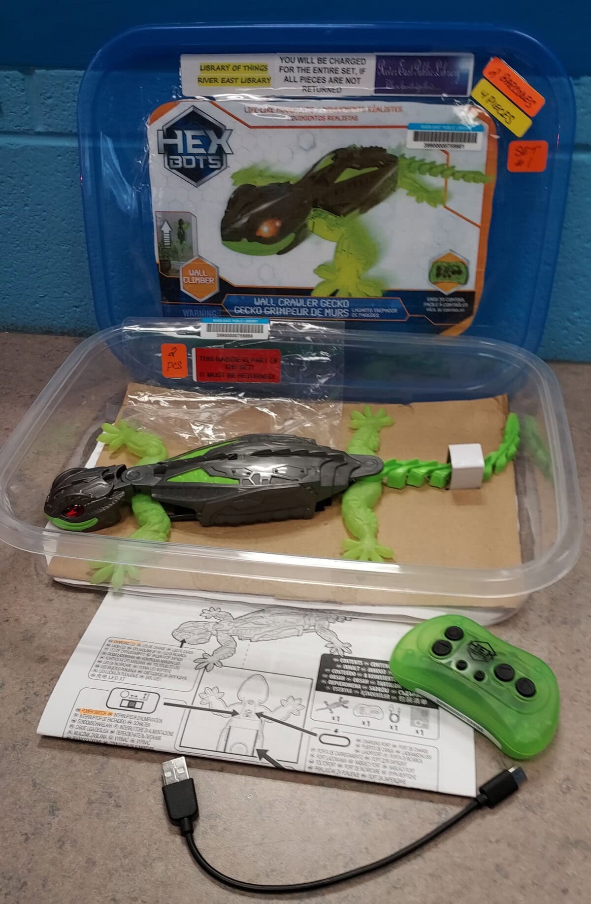 A HEXBUG remote control lizard toy is displayed in a clear plastic container with its packaging, green remote control, USB charging cable, and instruction sheet on a gray surface.