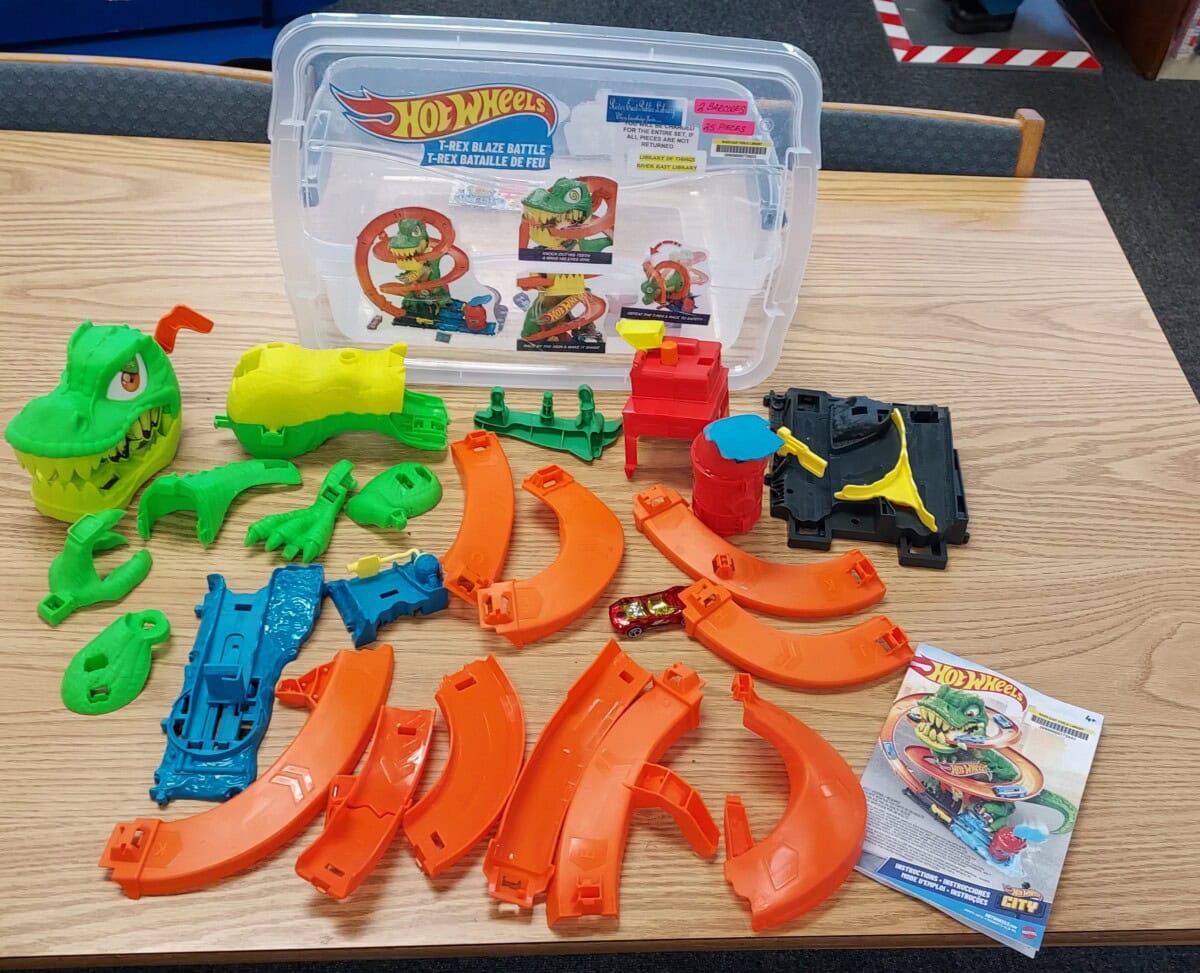 Hot Wheels playset parts are spread out on a table, including orange track pieces, green dinosaur parts, a small red car, and an instruction manual, with the packaging box displayed behind them.
