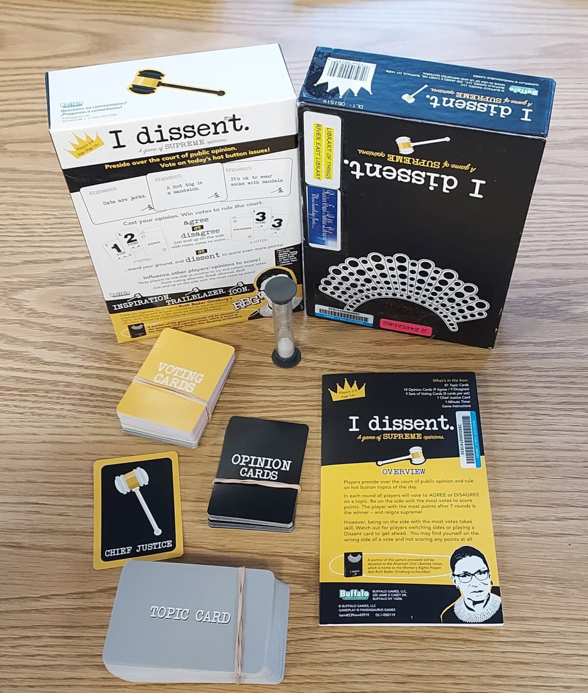 The board game I Dissent is displayed on a wooden surface. The game box, instruction sheet, cards labeled Voting, Opinion, Chief Justice, and Topic, along with a sand timer, are all visible.