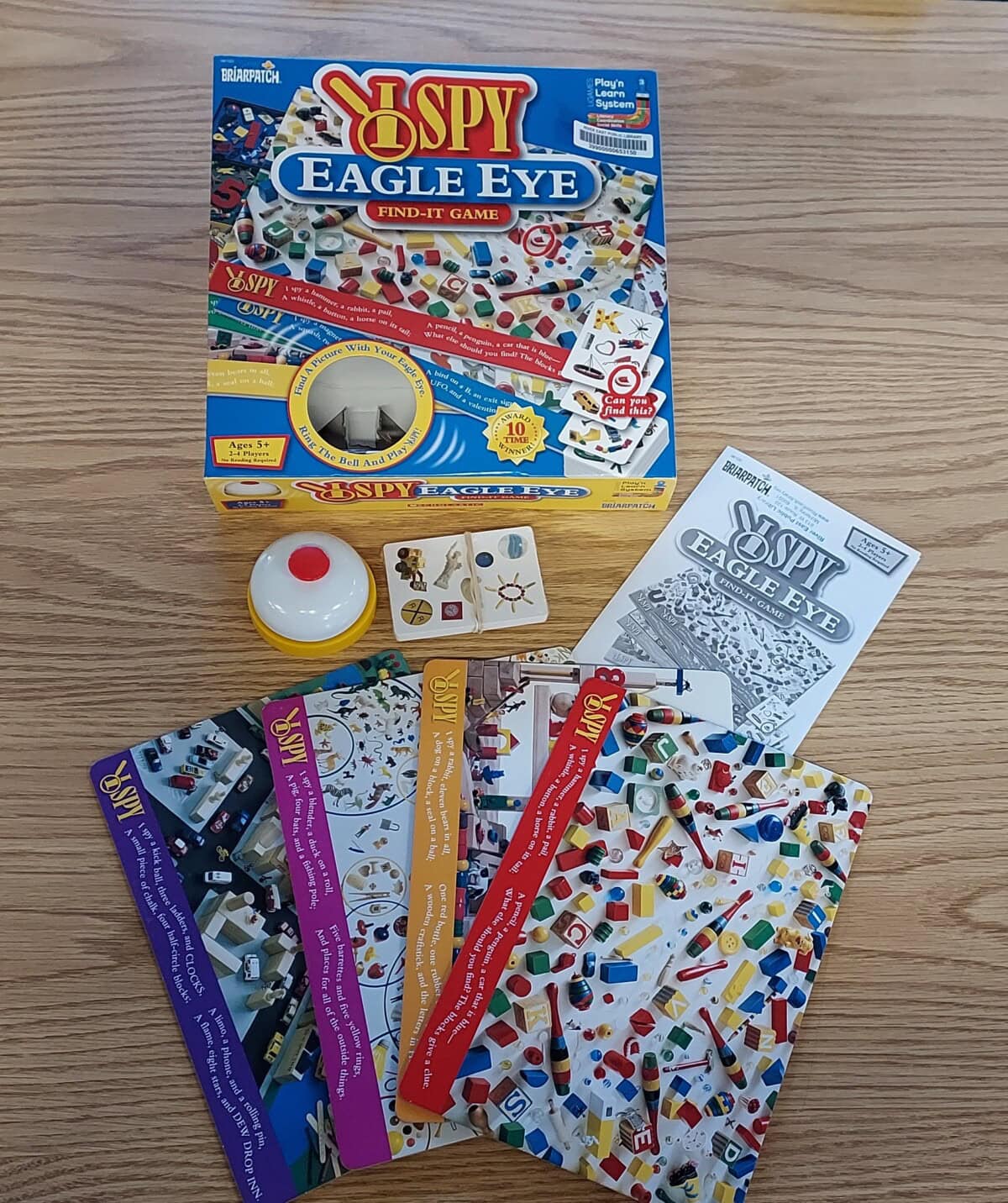 The image shows the I Spy Eagle Eye board game, including the game box, cards with colorful objects, a yellow and white buzzer, instruction booklet, and small square cards, all arranged on a wooden surface.