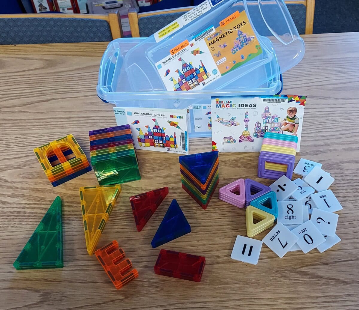 Colorful magnetic building tiles of various shapes are spread out on a wooden table alongside number cards, two instruction booklets, and a clear plastic storage box labeled “MAGNETIC TOYS.” Blue chairs are in the background.