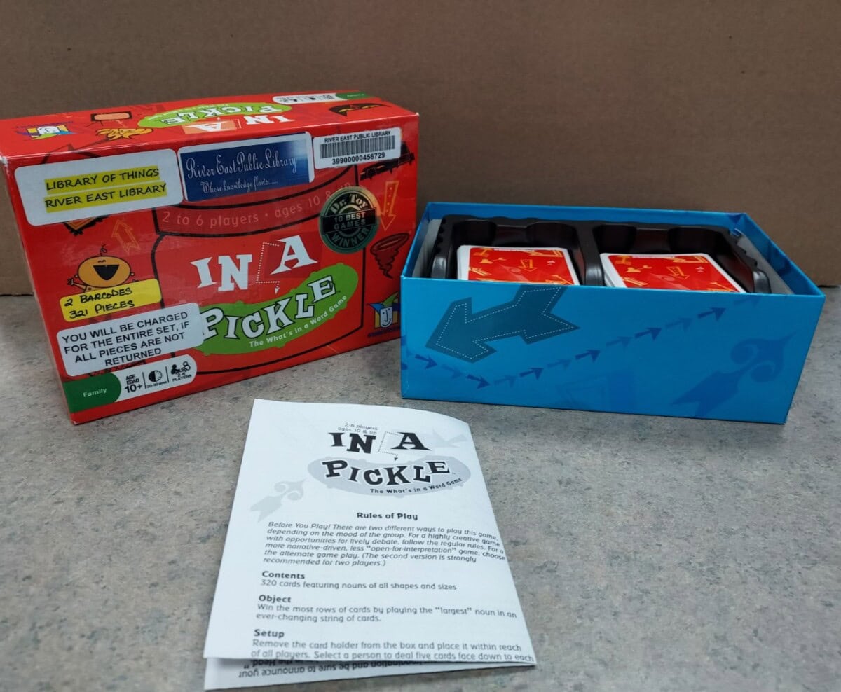 The image shows the “In a Pickle” card game with its box, a blue card tray containing stacks of cards, and a game instructions sheet laid out on a gray surface. The box features colorful graphics and labels.