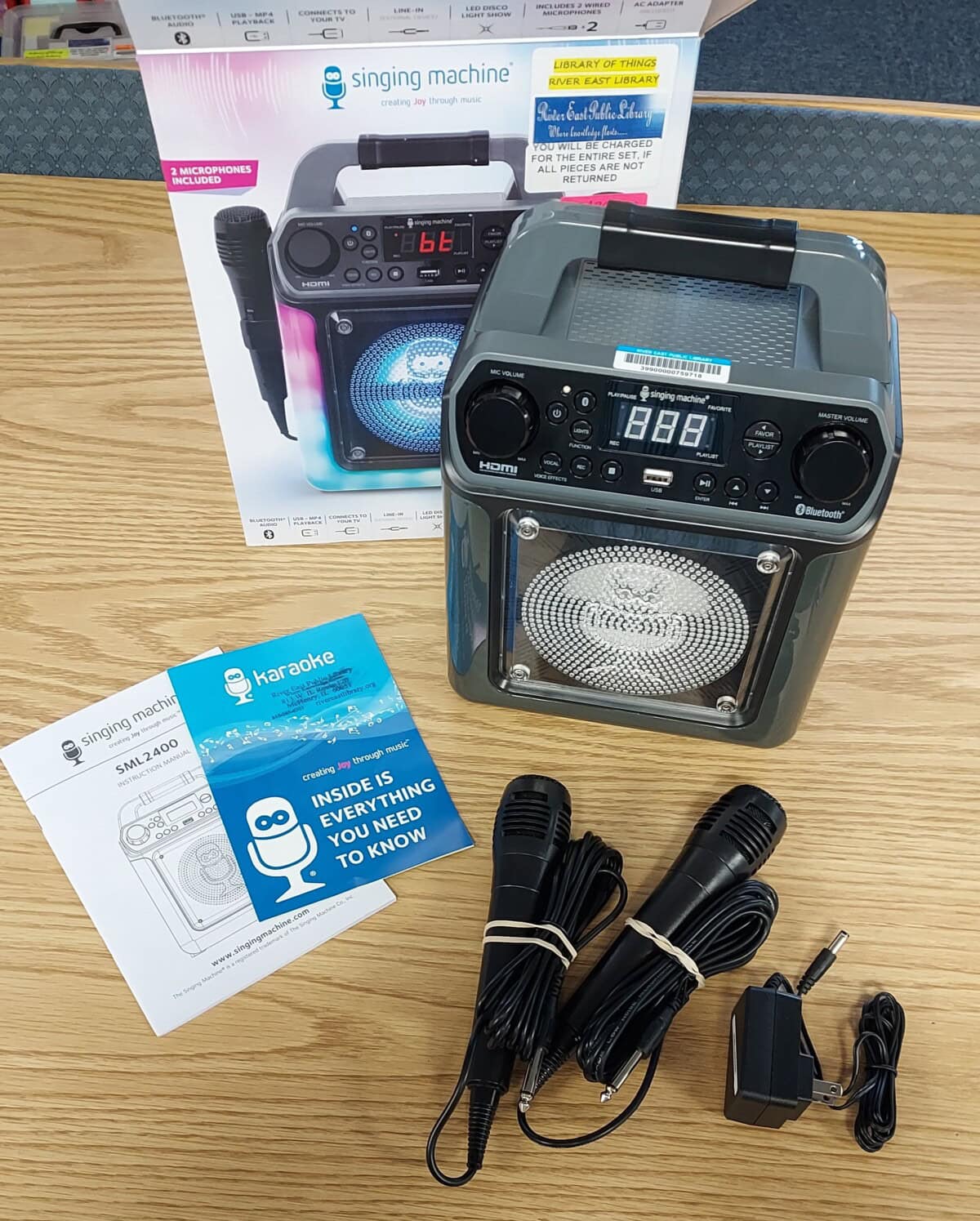 A karaoke machine set on a wooden table with its box, instruction manuals, two wired microphones, and a power adapter. The box features an image of the machine and the brand “Singing Machine.”.