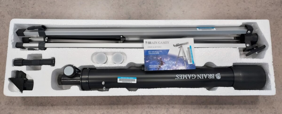 A boxed Brain Games telescope kit with a black telescope tube, tripod, eyepieces, and instructional booklet arranged in a white foam insert.
