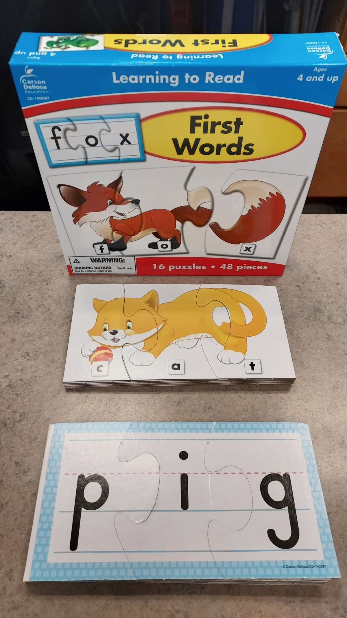 A box labeled First Words with a fox illustration, a cat puzzle spelling cat, and a separate puzzle spelling pig are displayed on a gray surface.