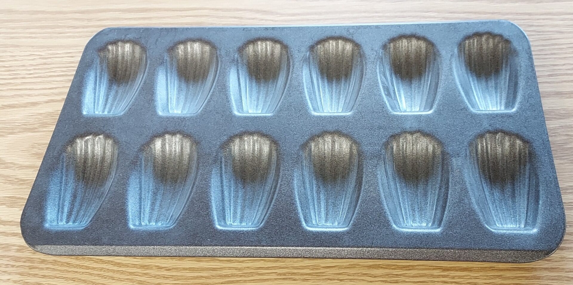 A metal madeleine baking tray with twelve shell-shaped molds is placed on a wooden surface.