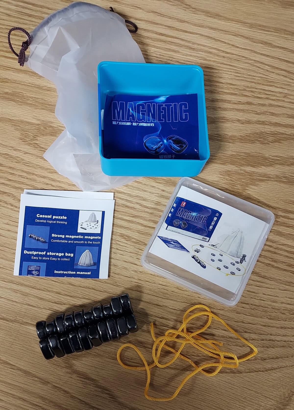 A blue plastic box labeled MAGNETIC sits on a wood surface with magnetic rings, instruction cards, a transparent case, a yellow string, and a white storage bag spread around it.