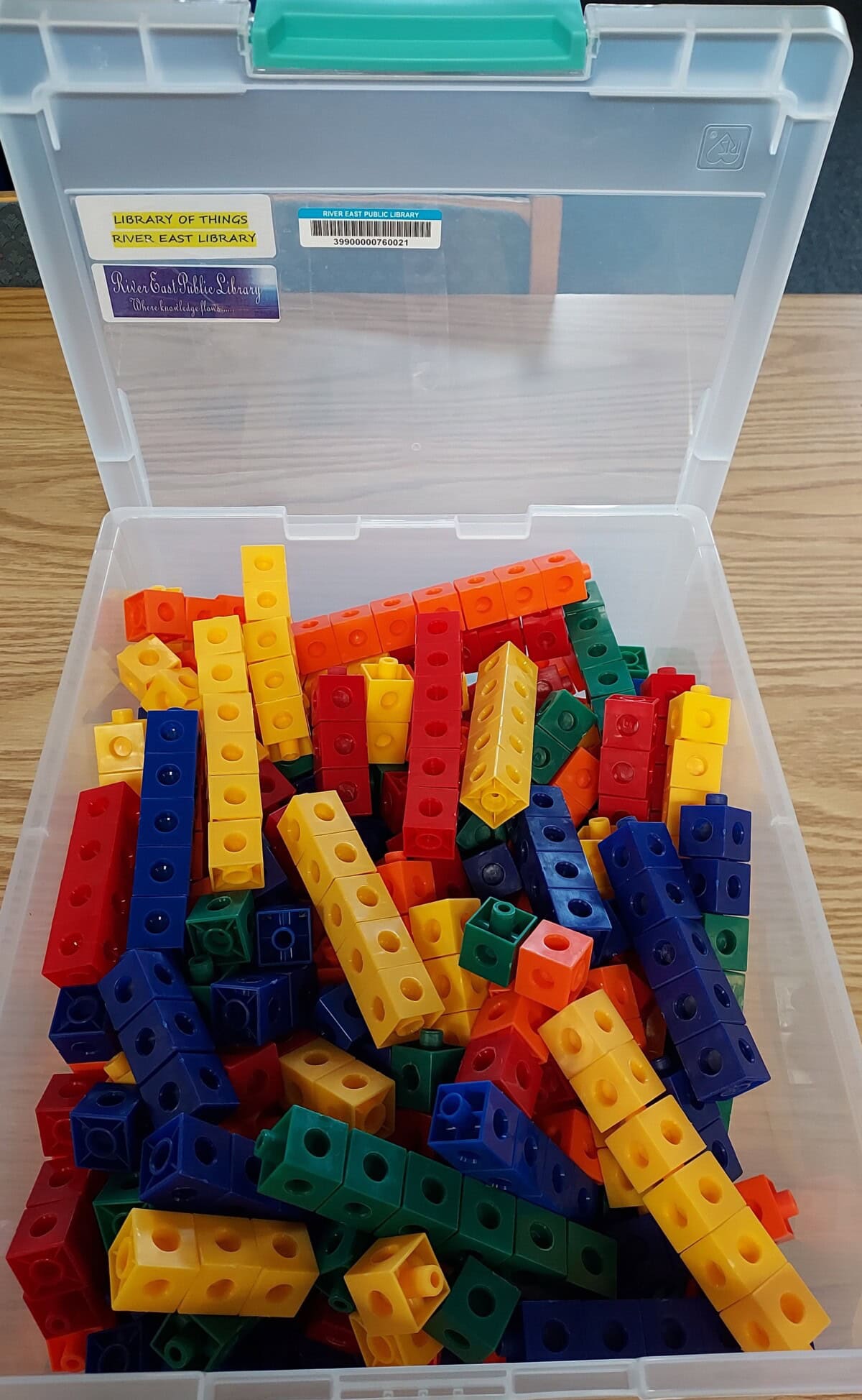 An open plastic box filled with colorful interlocking counting cubes in red, yellow, blue, green, and orange, sitting on a wooden surface. Labels for Library of Things and a library are on the box lid.