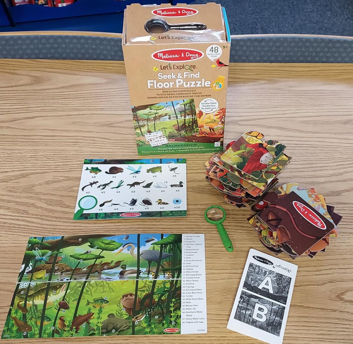A Melissa & Doug Seek & Find Floor Puzzle set is on a table, including a puzzle box, assembled puzzle, activity sheets, a green magnifying glass, and a stack of puzzle pieces with animal and nature illustrations.