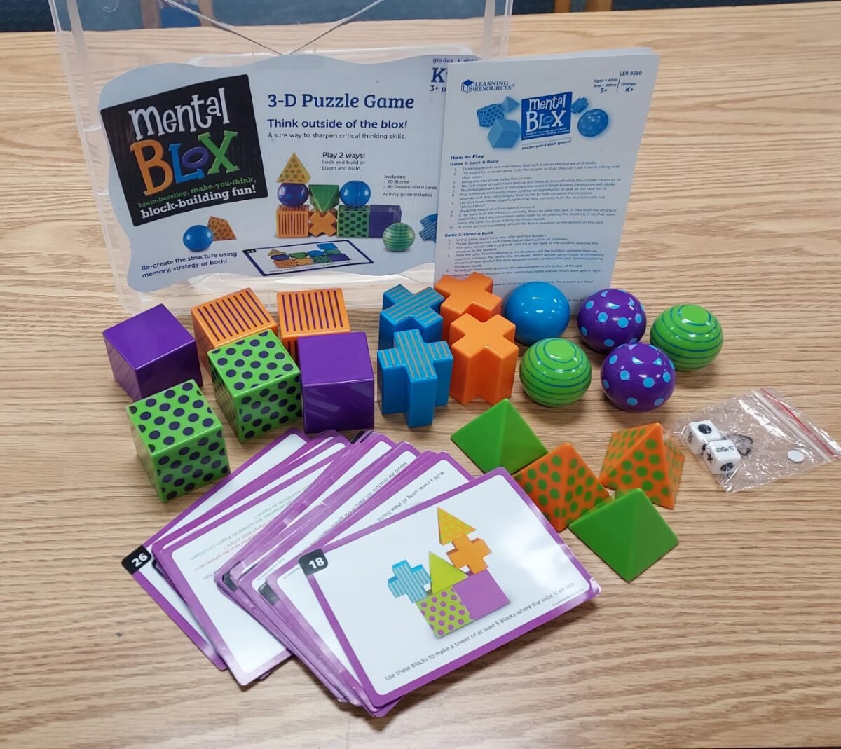 A set of colorful geometric blocks, game cards, dice, and instructions for the Mental Blox 3-D Puzzle Game is spread out on a wooden table. The blocks include cubes, spheres, pyramids, and crosses in various colors and patterns.