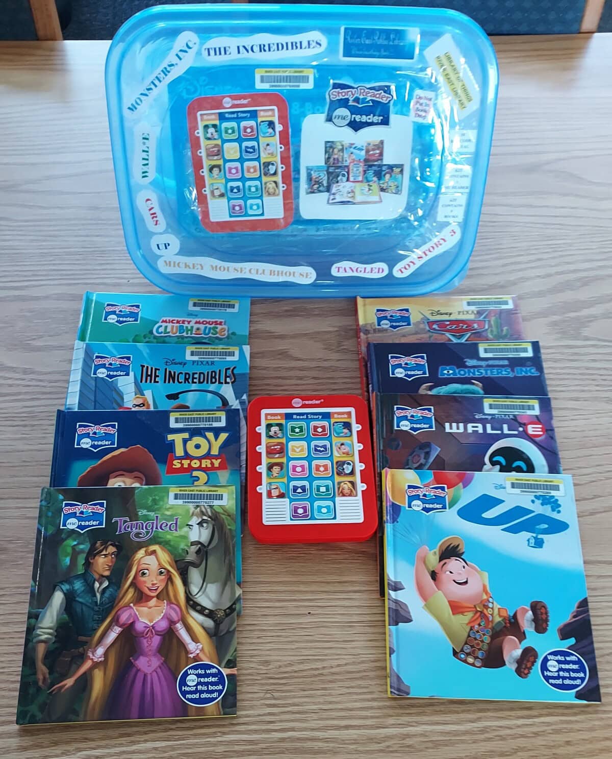 A set of Disney-themed childrens books and a red electronic Me Reader are displayed on a wooden table. Titles include Toy Story, Cars, Monsters Inc., WALL-E, UP, Tangled, and The Incredibles.