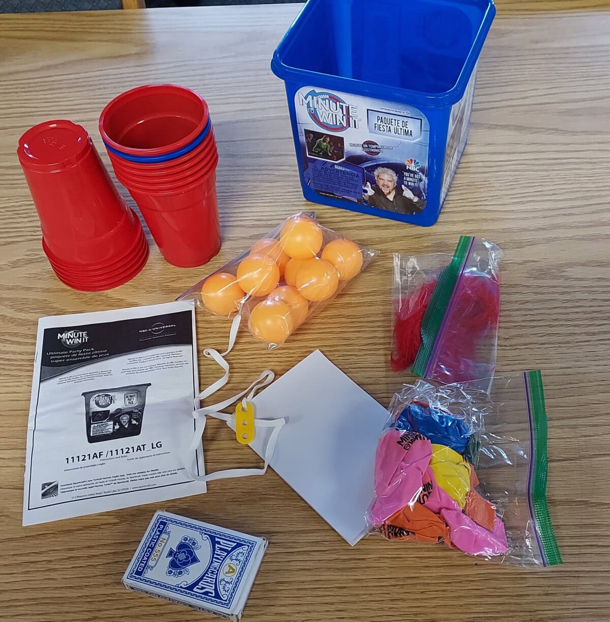 A collection of game supplies on a wooden table, including red plastic cups, orange ping pong balls, playing cards, balloons, feathers, a plastic bucket, string, instruction sheets, and a white card.
