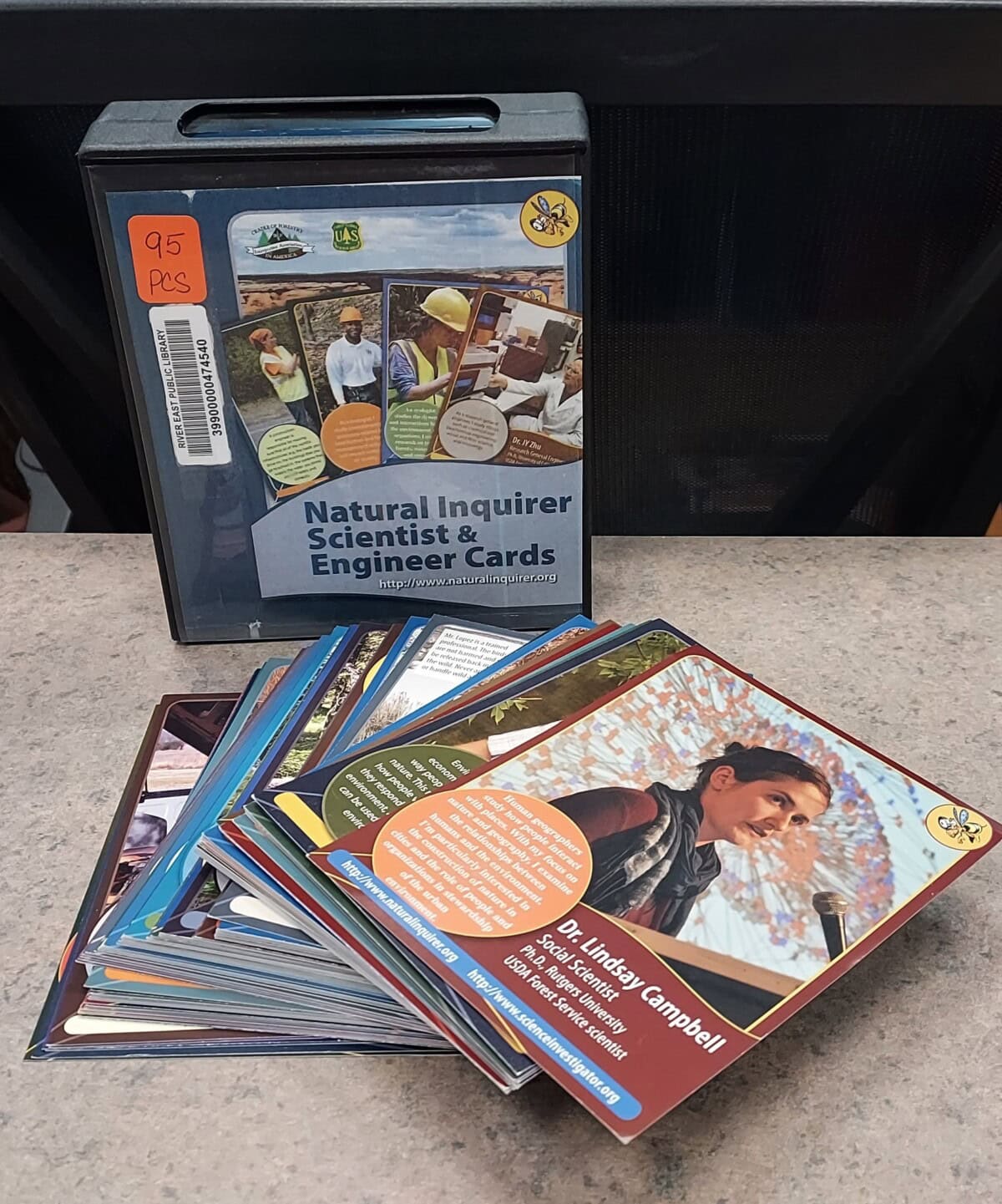 A binder labeled Natural Inquirer Scientist & Engineer Cards stands upright behind a spread of colorful trading cards on a countertop; one visible card features a photo and info about Dr. Lindsey Campbell.