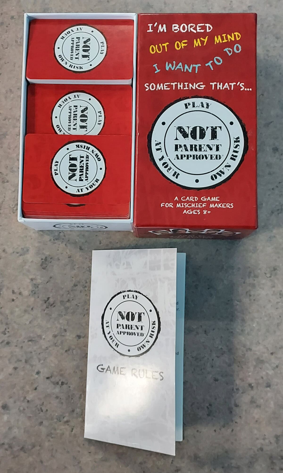 A card game box labeled Not Parent Approved sits open on a countertop. Red cards are stacked inside, and a booklet titled Game Rules lies in front of the box.