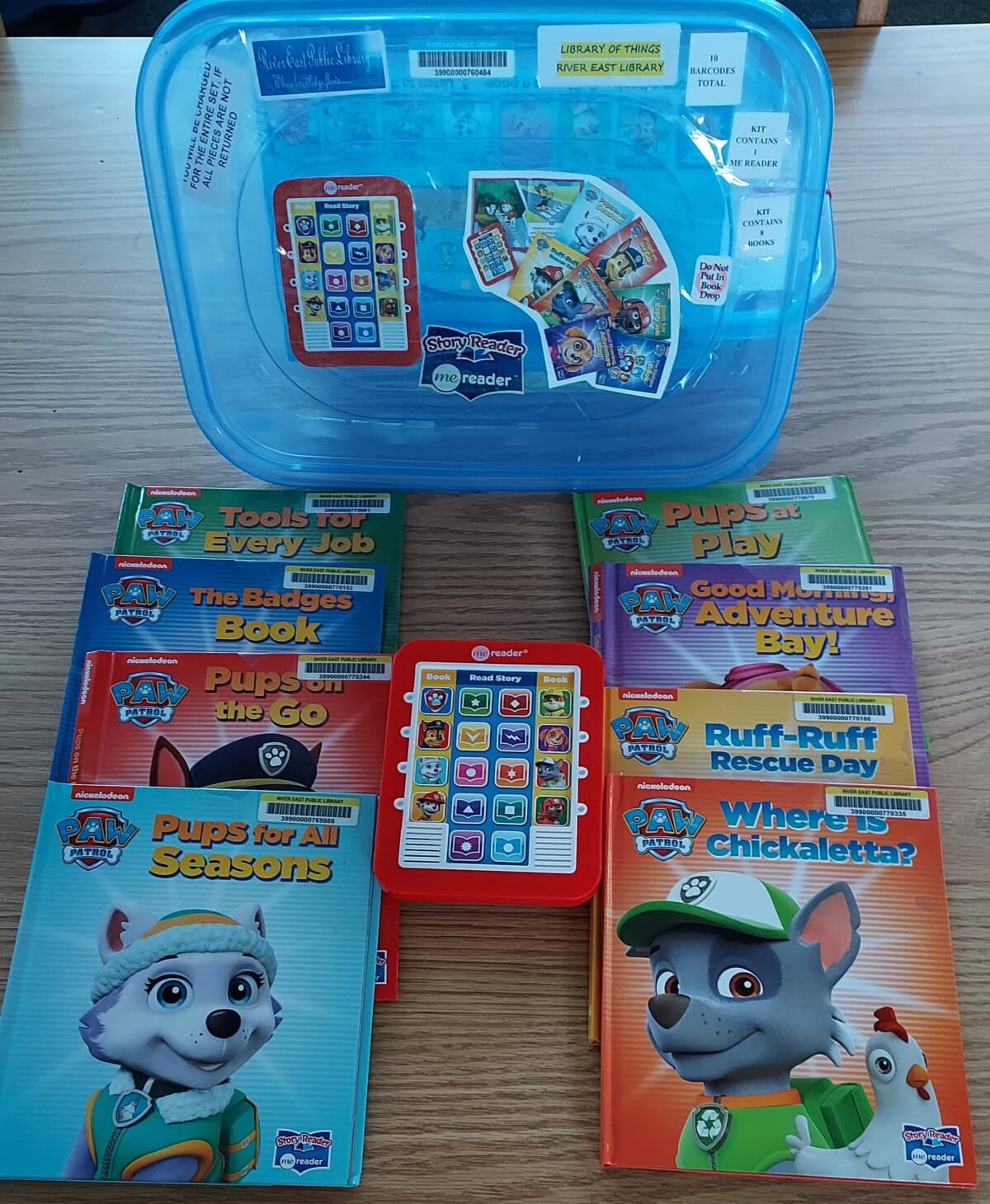 A blue plastic box labeled “Library of Things” with PAW Patrol books, a red and white interactive Me Reader device, and several colorful children’s books arranged on a wooden surface.