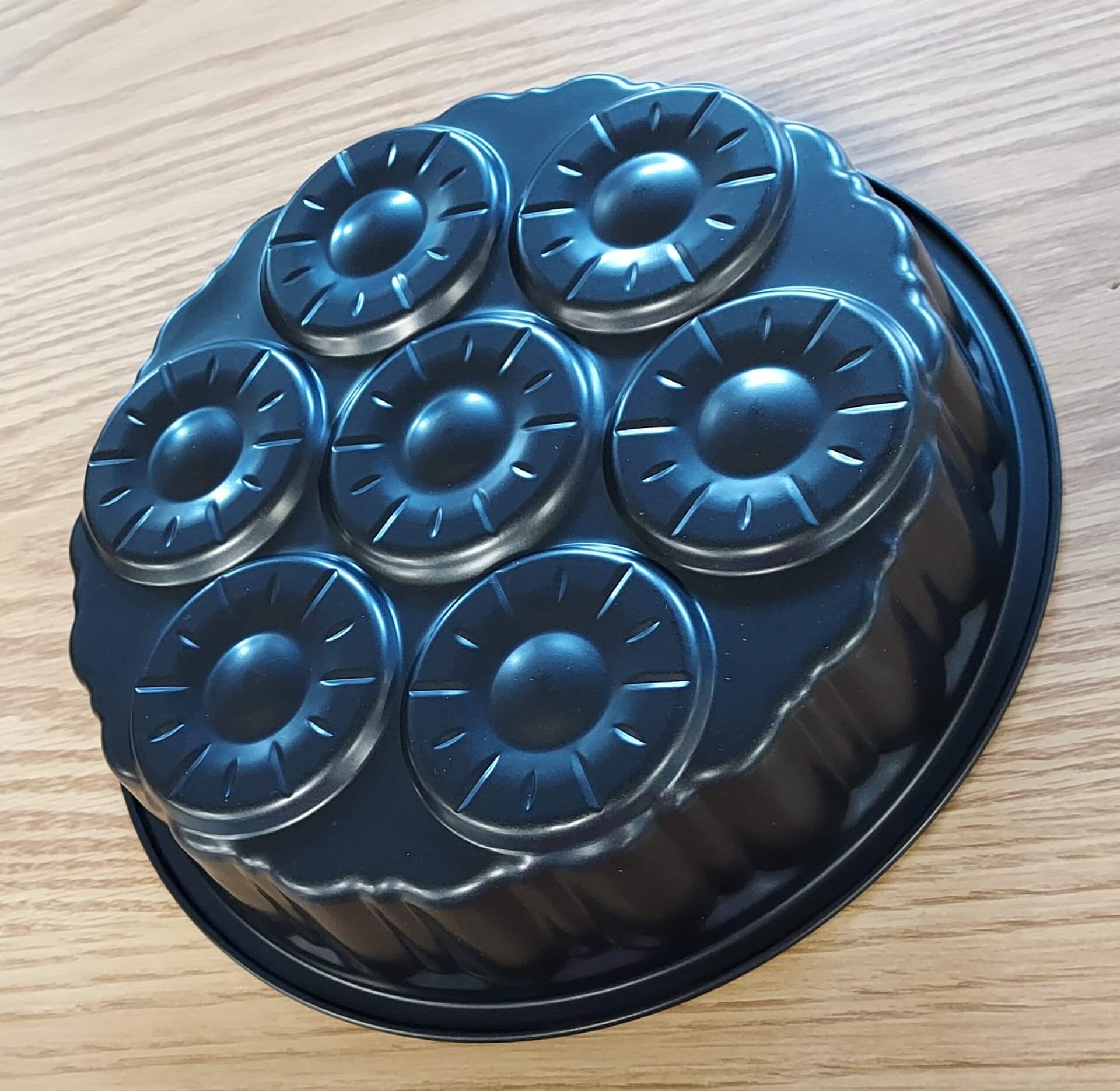 A black metal baking tin with seven round, fluted molds arranged in a circle, sitting on a light wood surface.