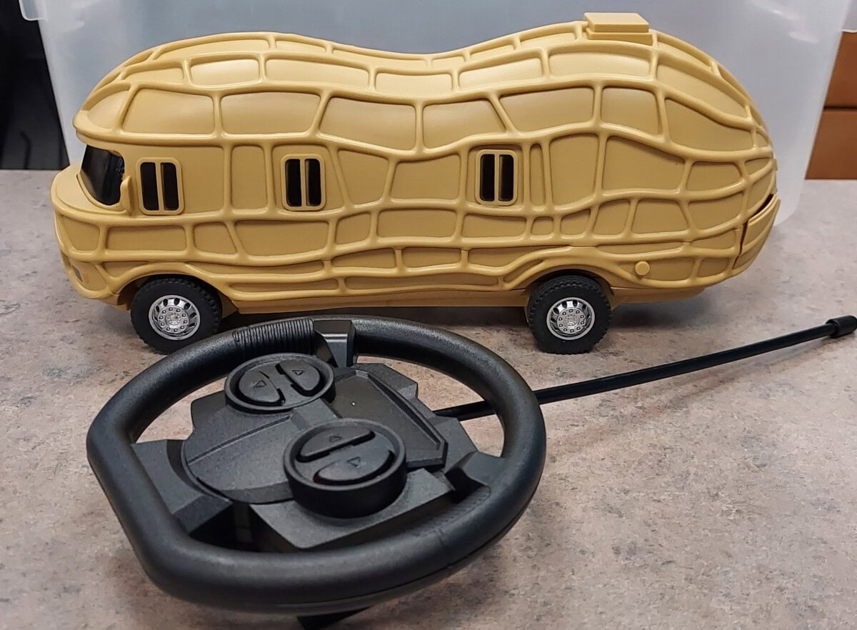 A toy vehicle shaped like a peanut with windows and wheels sits on a table next to a black remote control shaped like a steering wheel.