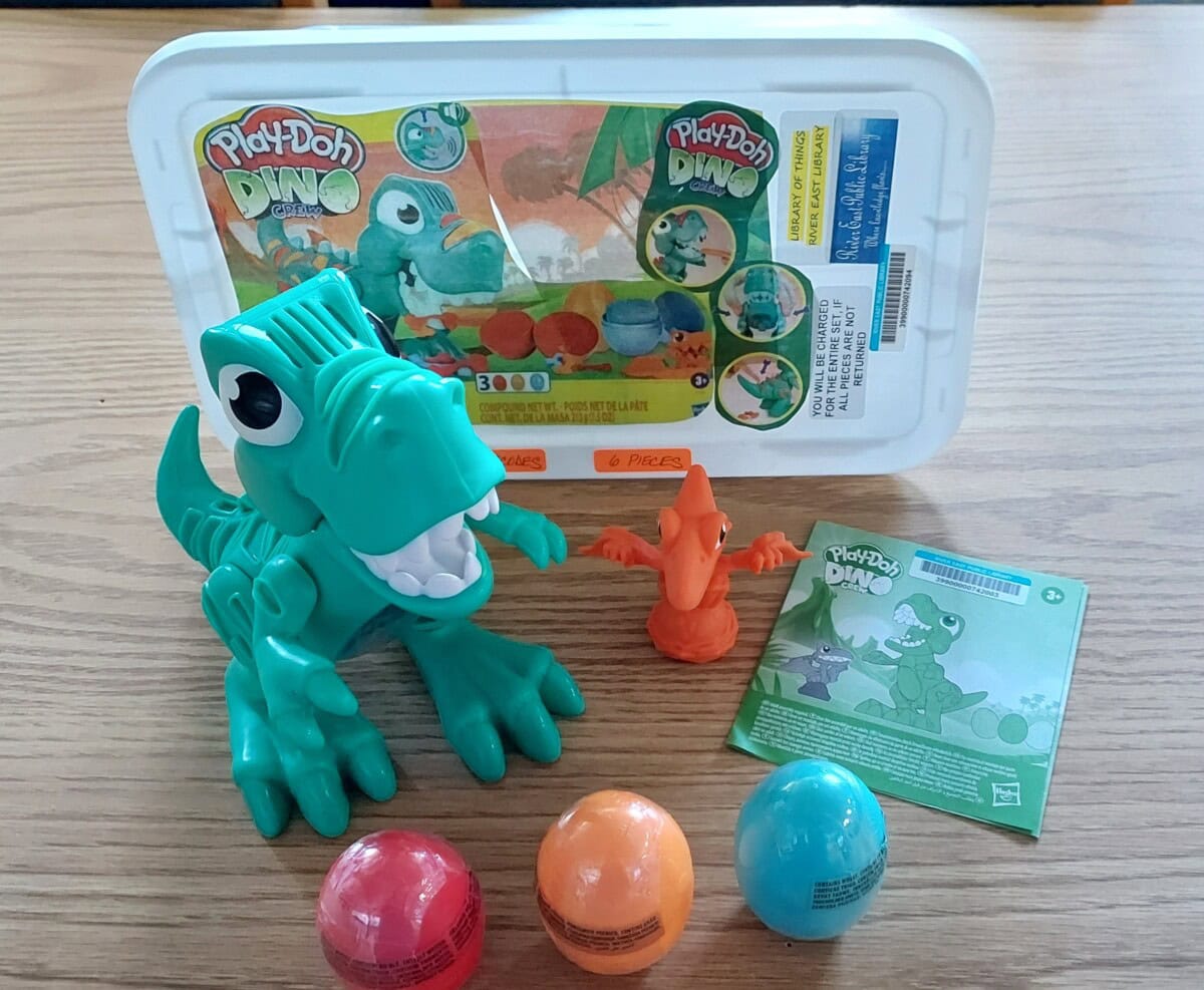 A Play-Doh Dino set with a green dinosaur toy, a small orange dinosaur figure, three colorful Play-Doh containers, and an instruction booklet on a wooden surface. The sets packaging is visible in the background.