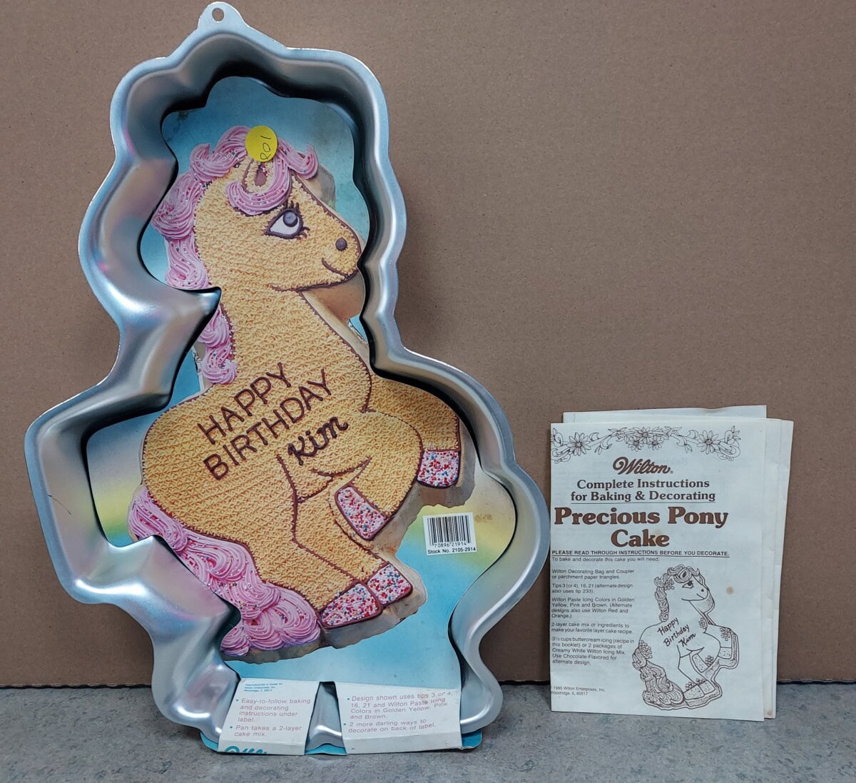 A Wilton pony-shaped cake pan with a decorated example cake reading Happy Birthday Kim, next to an instruction booklet titled Precious Pony Cake showing a line drawing of the pony cake.