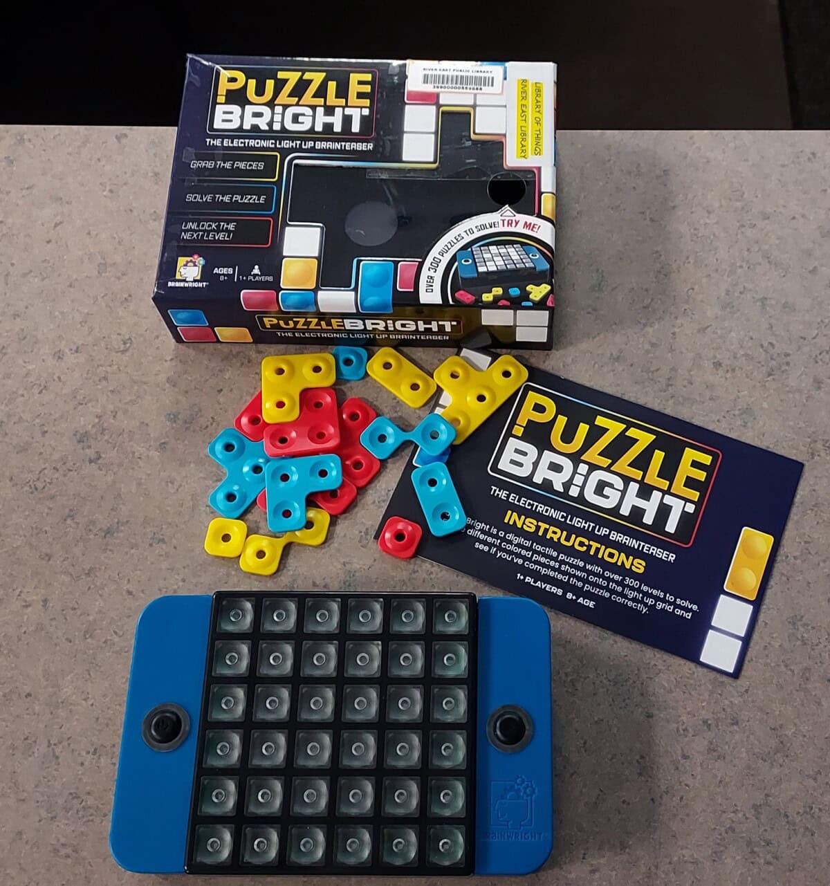 A PuzzleBright electronic light-up game sits on a table with its box, colorful yellow, red, and blue plastic pieces, and an instruction booklet scattered around it.