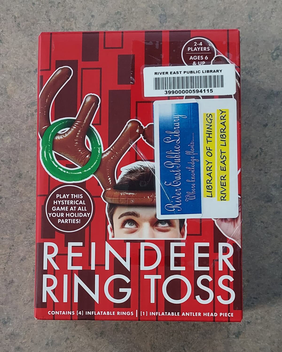 A boxed game called Reindeer Ring Toss shows a person wearing inflatable antlers with rings in mid-air above them. Library barcodes and labels are on the box. For 2-4 players, ages 6 and up.