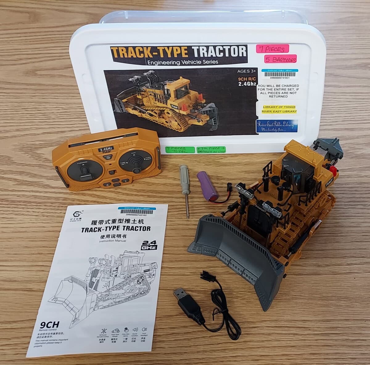A remote-control construction bulldozer toy set with yellow bulldozer, orange remote, screwdriver, battery, USB charger, instruction manual, and labeled storage box on a wooden surface.