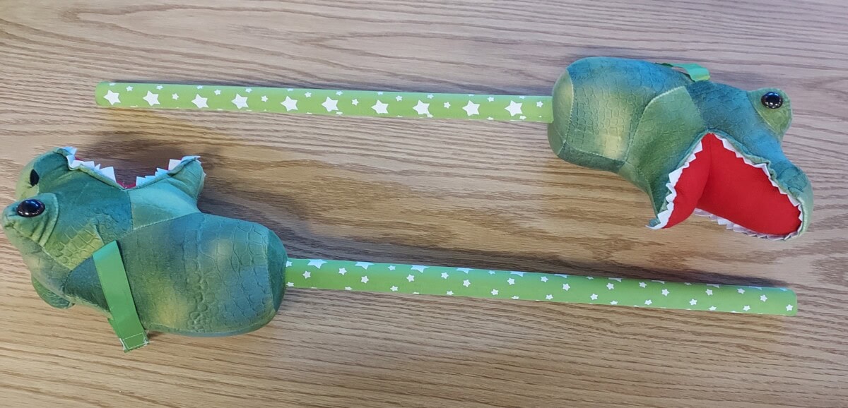 Two green toy grabbers with dinosaur heads and star-patterned handles lie on a wooden surface. The dinosaur mouths are open, showing red inside.