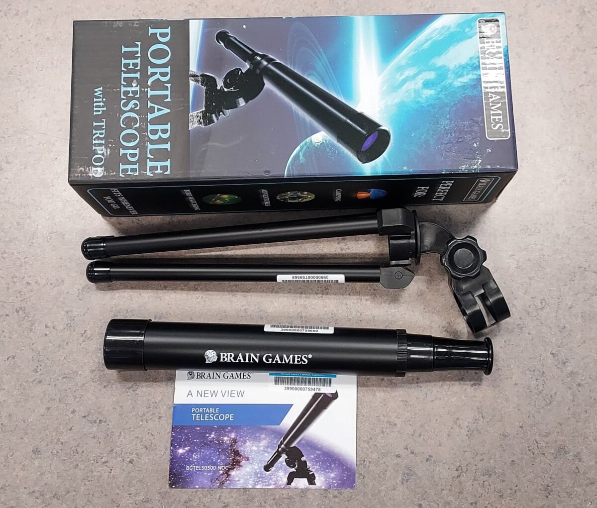 A portable telescope set from Brain Games is shown with its box, two detachable tripod legs, and an instruction manual on a tabletop. The box features an image of the telescope and space graphics.
