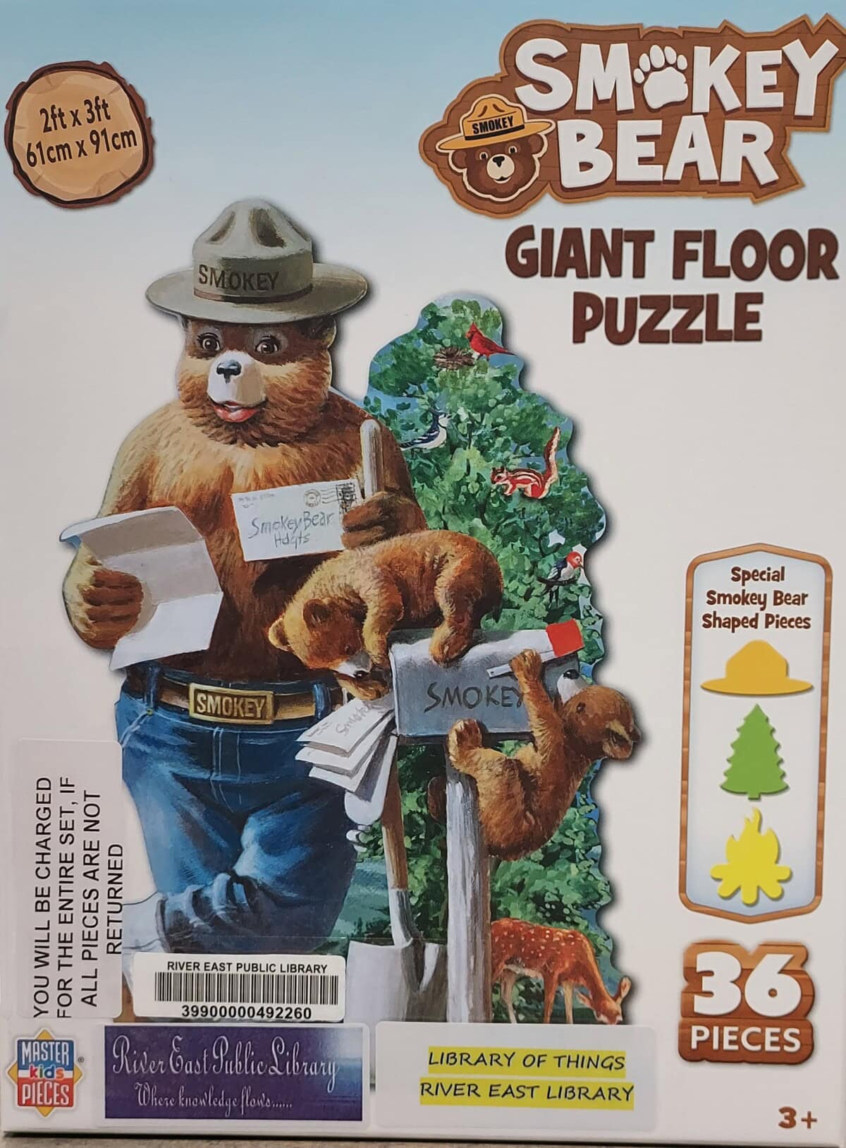Box cover of a 36-piece Smokey Bear giant floor puzzle, showing Smokey Bear holding a shovel and reading mail. A sign reads Library of Things, River East Public Library. For ages 3 and up.