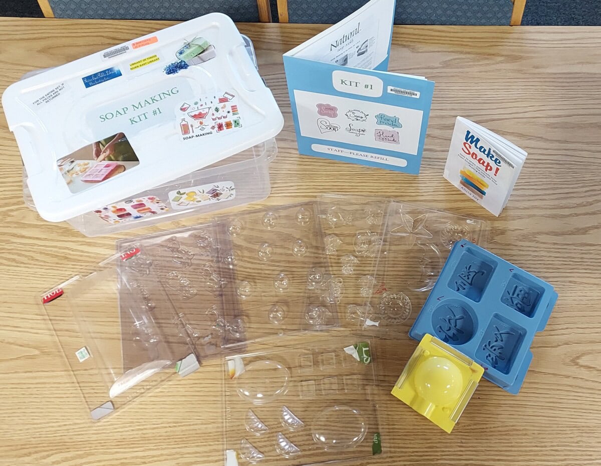 A soap making kit on a wooden table includes a plastic storage box, several soap molds in clear and blue plastic, instructional booklets, and soap making guides.