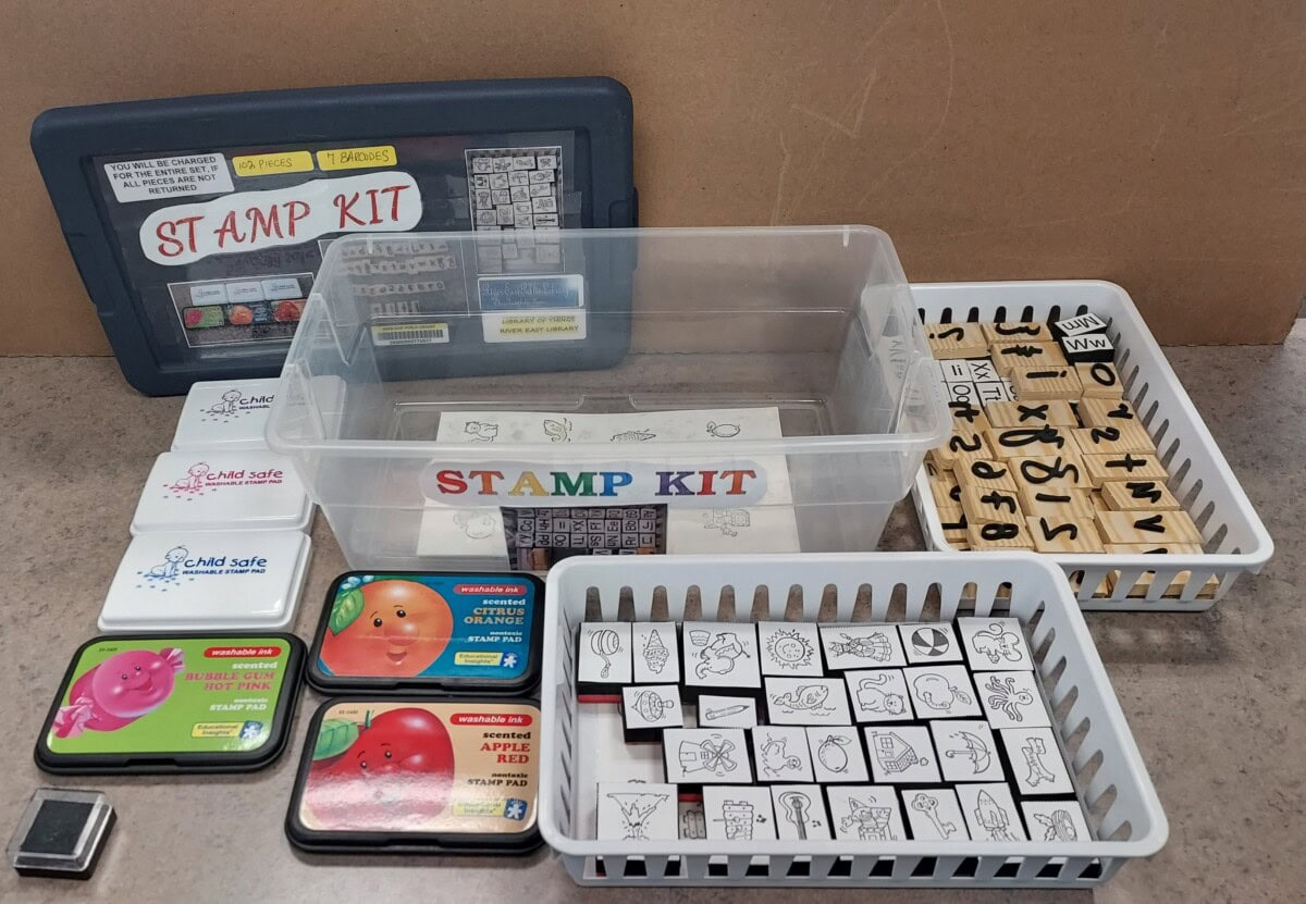 A stamp kit with various rubber stamps, ink pads, and plastic trays is displayed on a table. The items include a labeled box, assorted stamp designs, and colorful ink pads in tins and plastic cases.