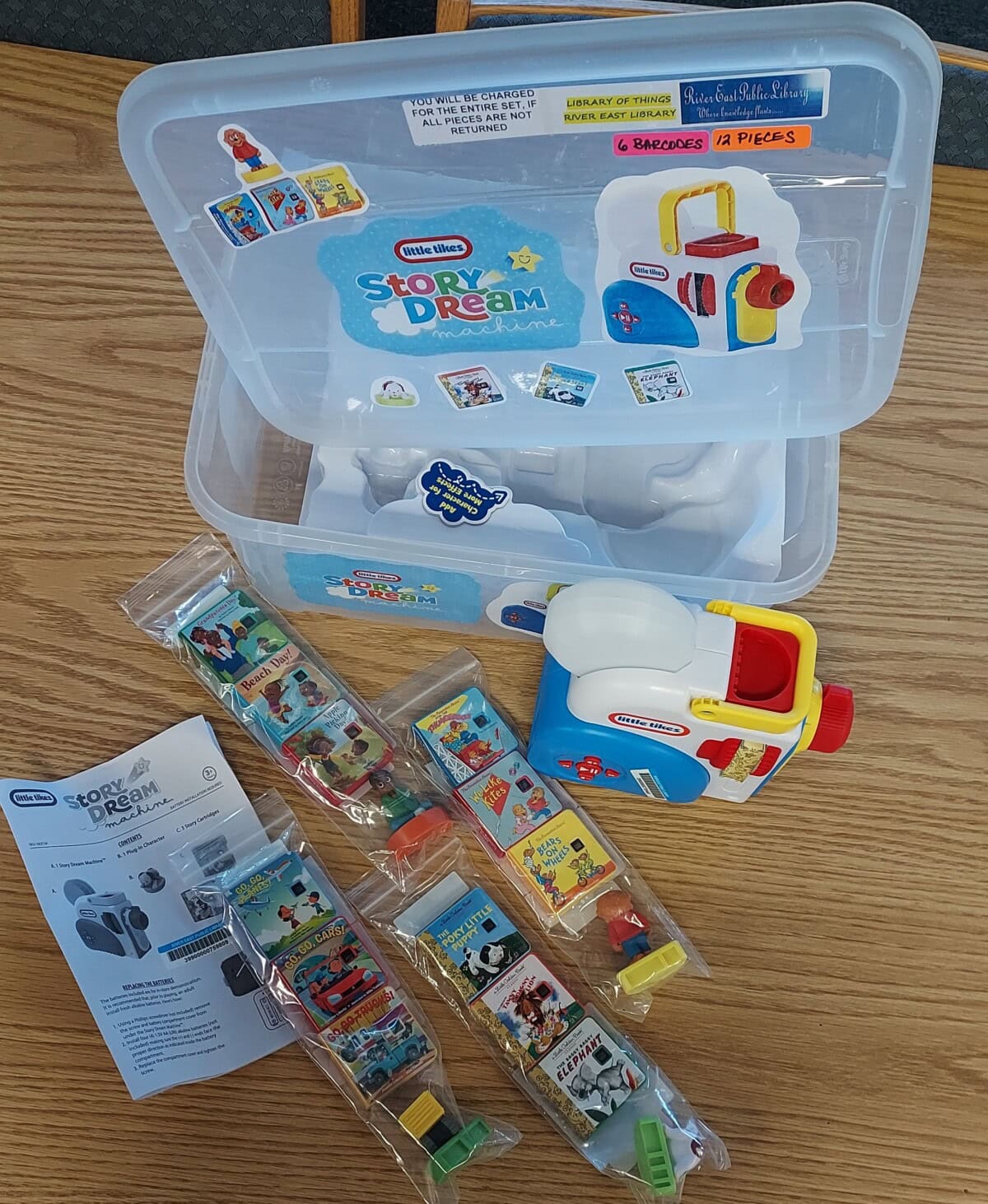 A Little Tikes Story Dream Machine set on a wooden table with an open plastic storage case, instruction manual, toy projector, and several film reels in clear packaging.