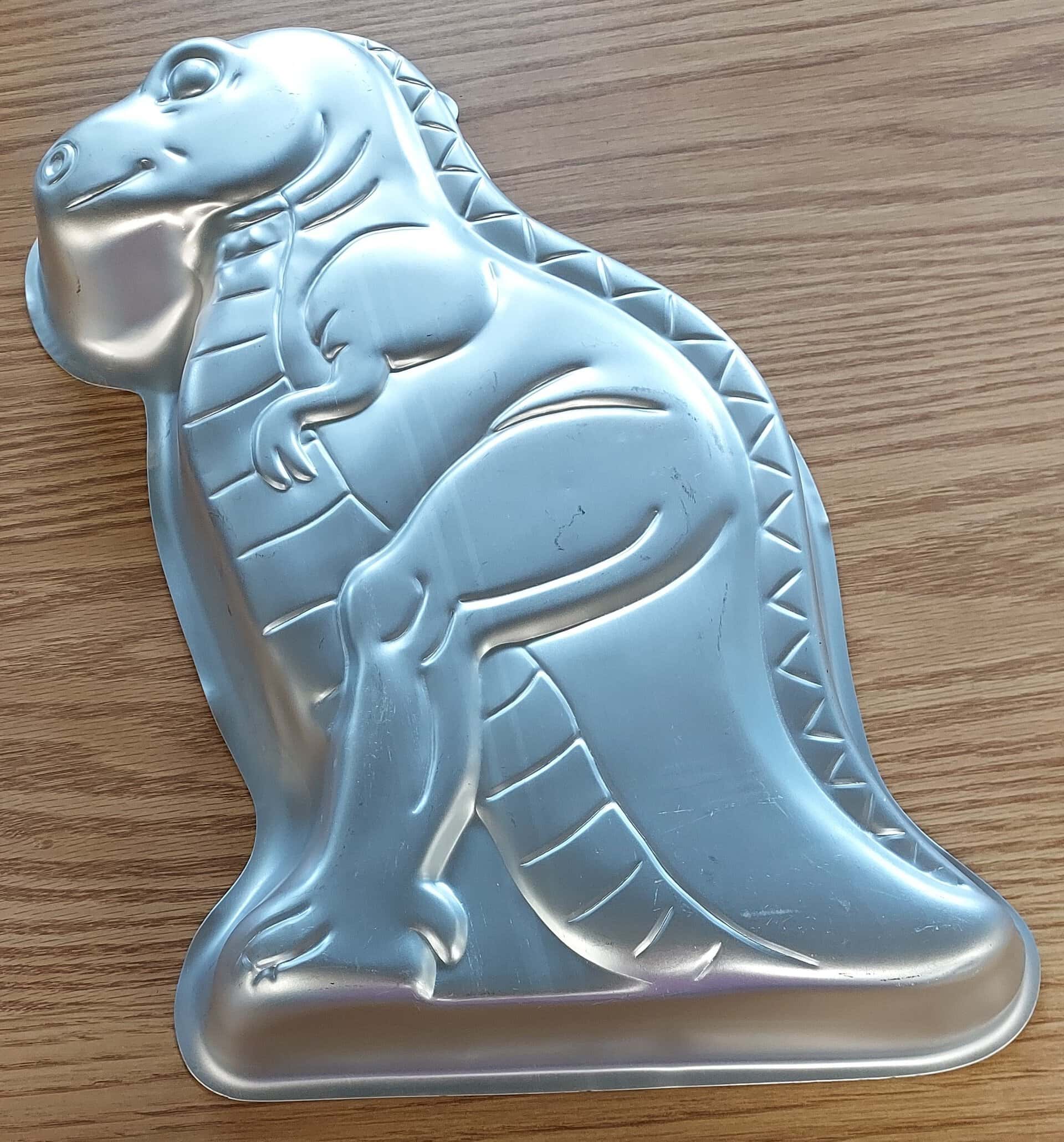 A silver metal cake pan shaped like a cartoon-style dinosaur, lying on a wooden surface. The dinosaur has a rounded snout, small arms, and a textured back.