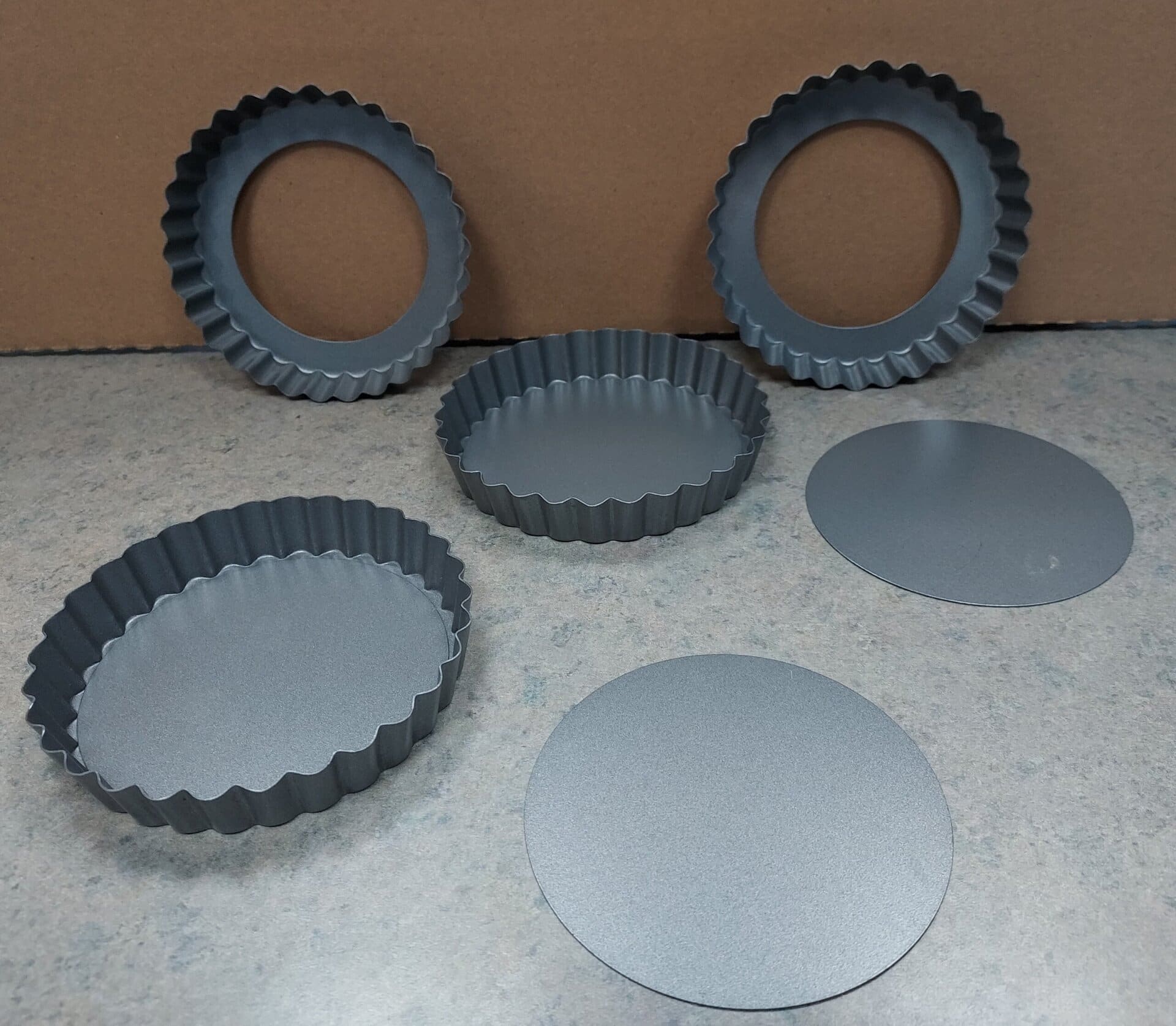 Five metal tart pans with removable bottoms are displayed on a countertop. Three round pan rings and two flat bases are separated and arranged in front of a plain brown background.