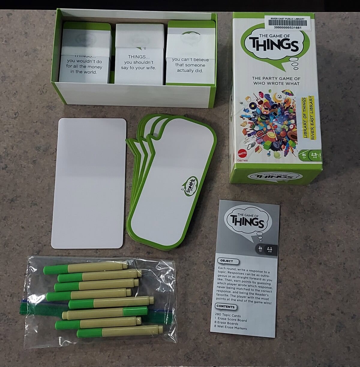 The image shows the board game The Game of Things opened on a table. Visible are cards, answer sheets, green-and-beige pens, a rule sheet, and the game box with its lid off, displaying the contents.