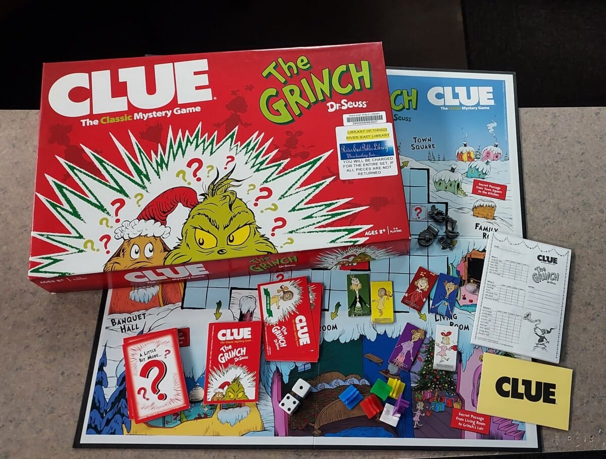 A Clue: The Grinch board game is set up on a table, showing the game board, cards, playing pieces, dice, a score sheet, and the game box featuring Dr. Seuss’s Grinch in festive holiday colors.