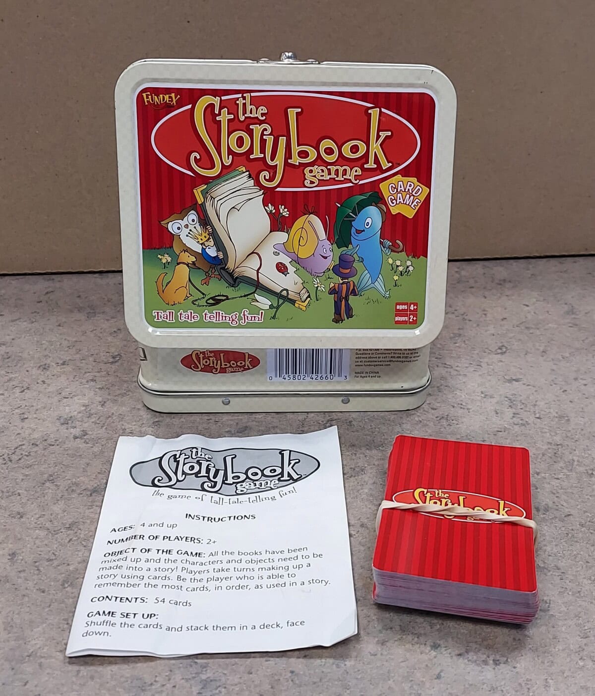 A tin box labeled The Storybook Game with cartoon book characters, a stack of red playing cards, and an instruction sheet are displayed on a countertop.