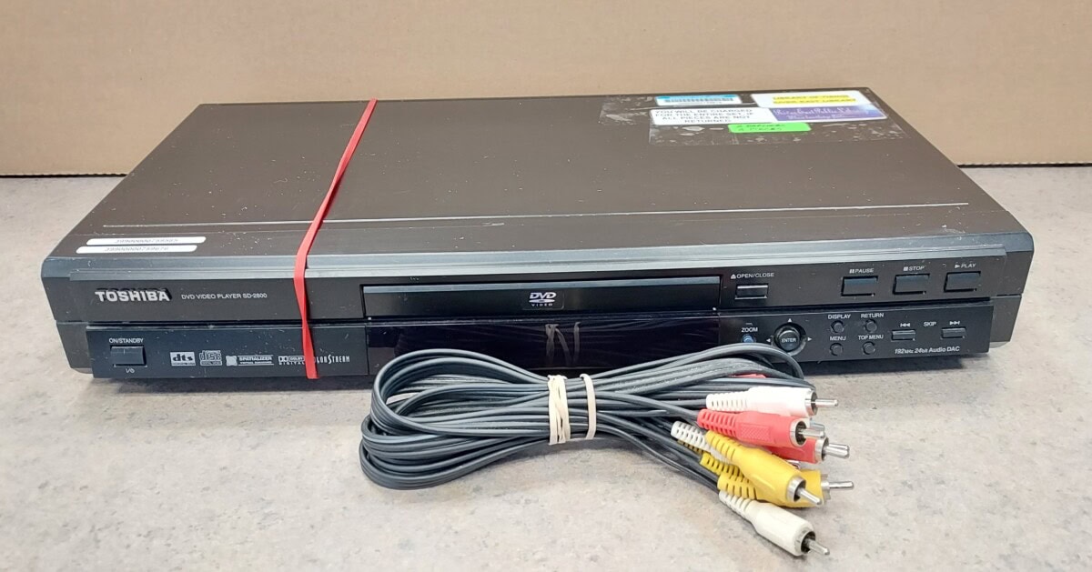 A Toshiba DVD video player sits on a counter with a red rubber band around it. In front of the player is a bundled AV cable with red, white, and yellow connectors. Several stickers are on the top of the player.