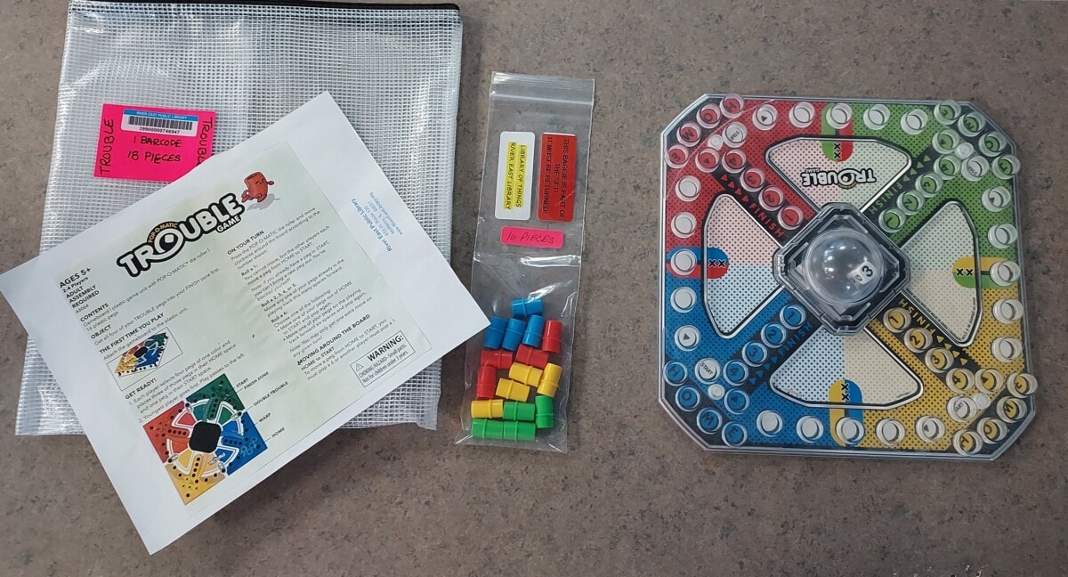 A Trouble board game set on a table, including the game board with pop-o-matic die, colored playing pieces in a bag, game instructions, and a mesh storage pouch with pink labels.