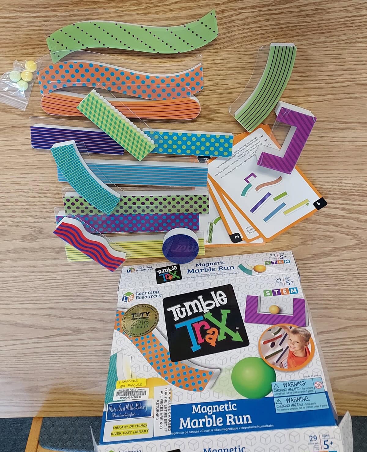 A Tumble Trax Magnetic Marble Run set on a wooden surface, showing colorful magnetic track pieces, marbles, instruction cards, and the product box with the set’s branding and illustrations.