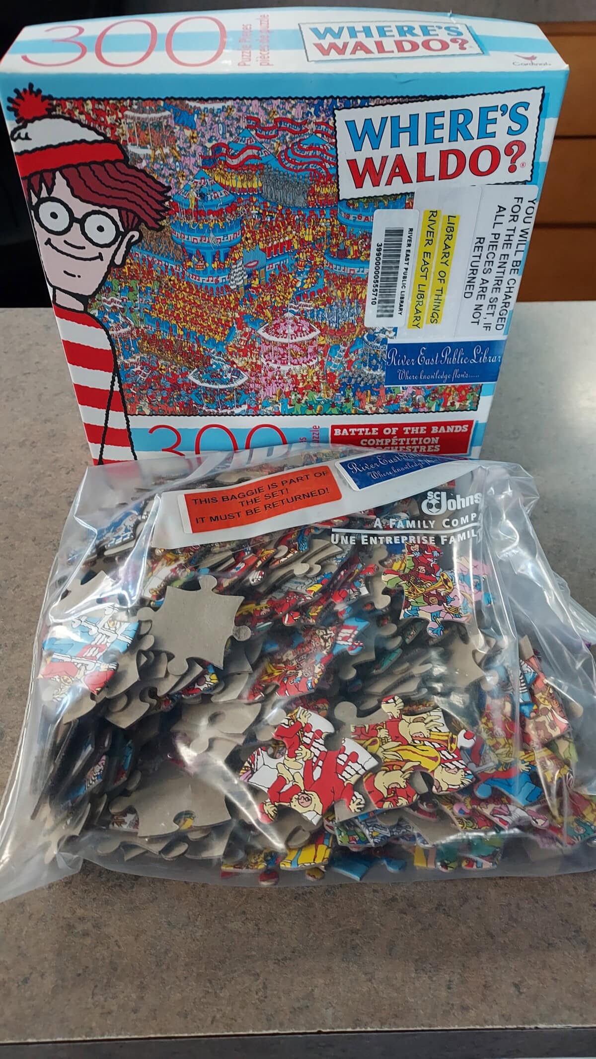 A 300-piece Wheres Waldo? puzzle box sits behind a clear plastic bag filled with loose puzzle pieces on a countertop. The puzzle features a crowded, colorful beach scene and Waldo’s cartoon image on the box.