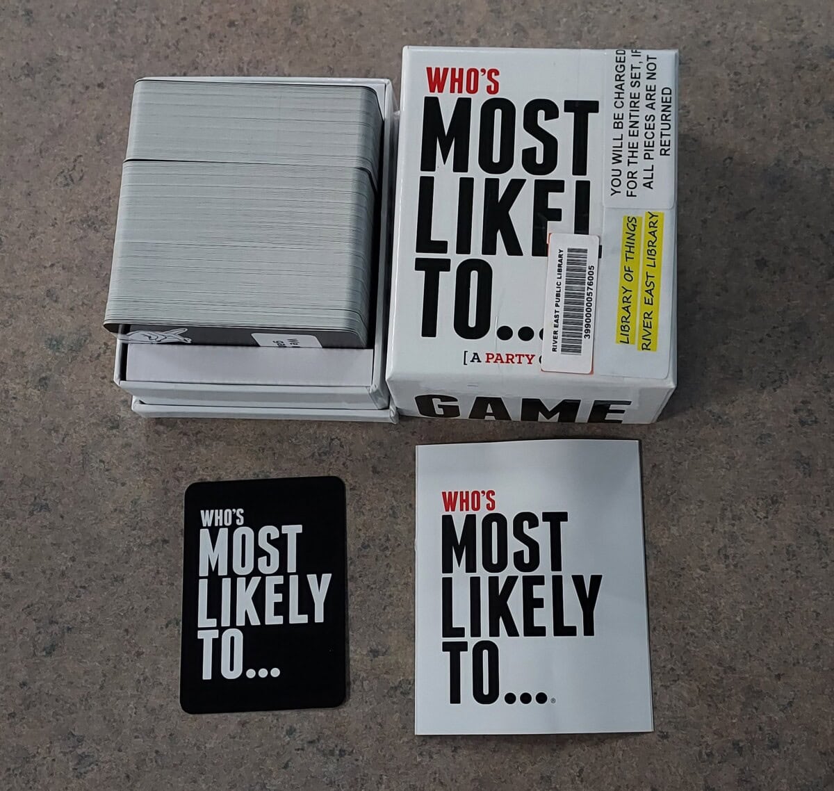 A card game titled Whos Most Likely To... is shown with its box, a stack of cards, and two cards laid out displaying the games title on a speckled surface.