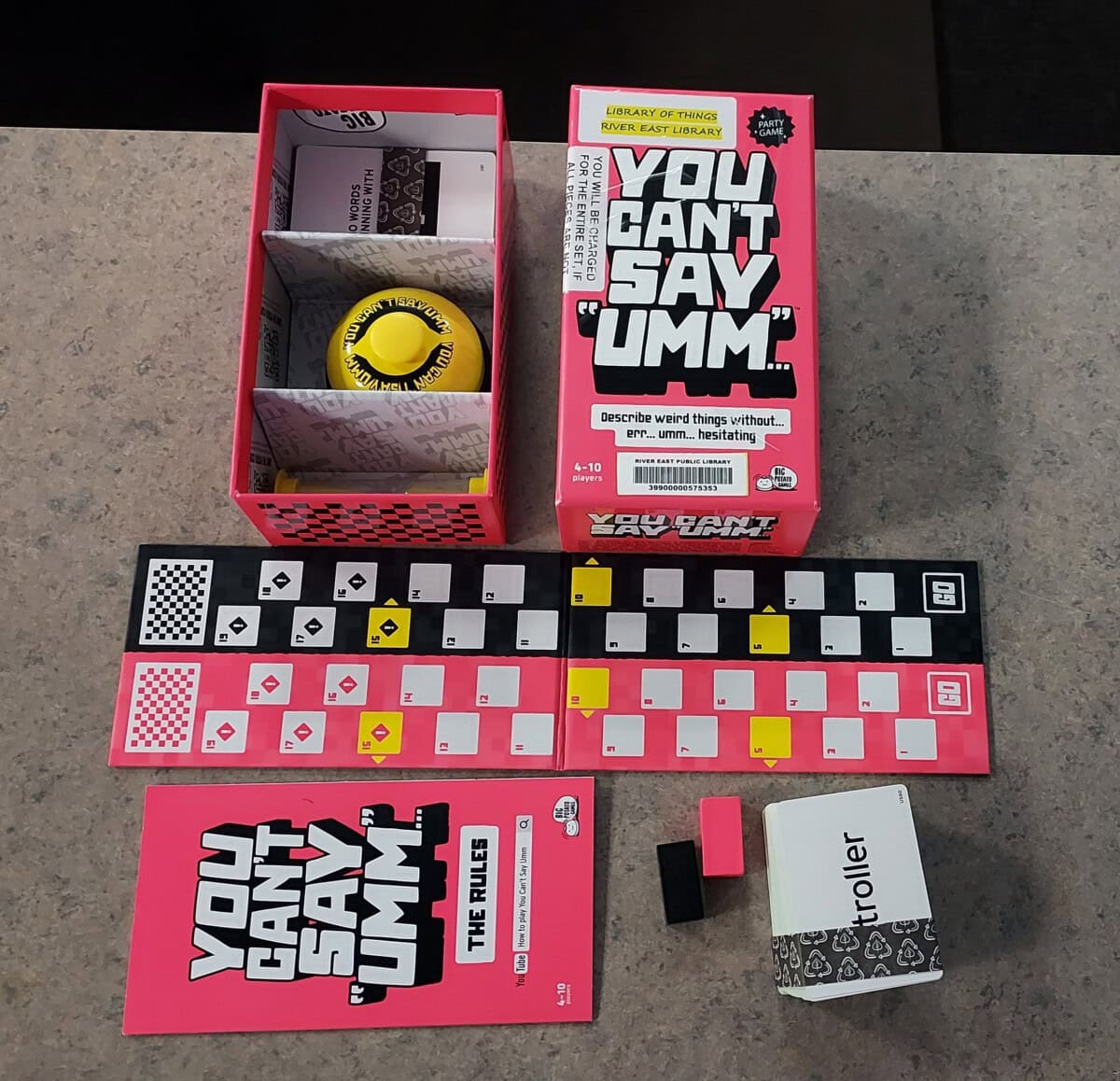 A board game called You Cant Say Umm is displayed on a table. The games box, rulebook, game board, cards, buzzer, and other components are neatly arranged in a flat lay style.