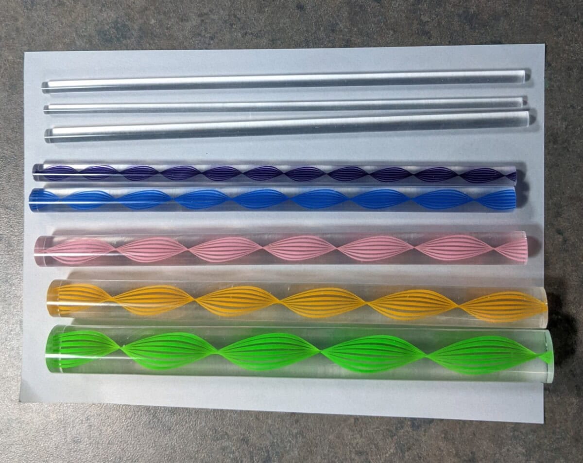 Seven glass stirring rods on paper: two clear, one blue, one purple, one pink, one yellow, and one green. The colored rods have twisted, spiral designs inside. They are arranged in parallel rows on a gray surface.