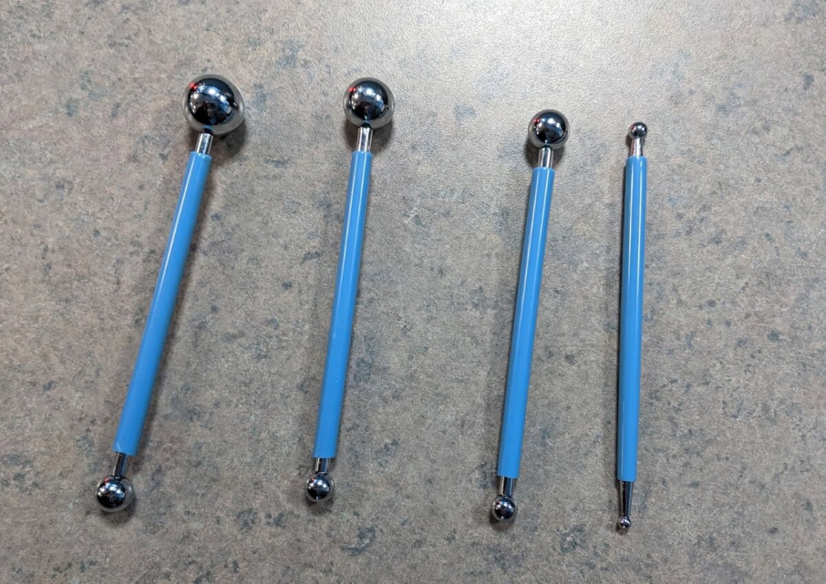 Four blue-handled metal ball stylus tools of varying ball sizes are arranged in a row on a speckled countertop. Each tool has a metallic ball tip on each end.