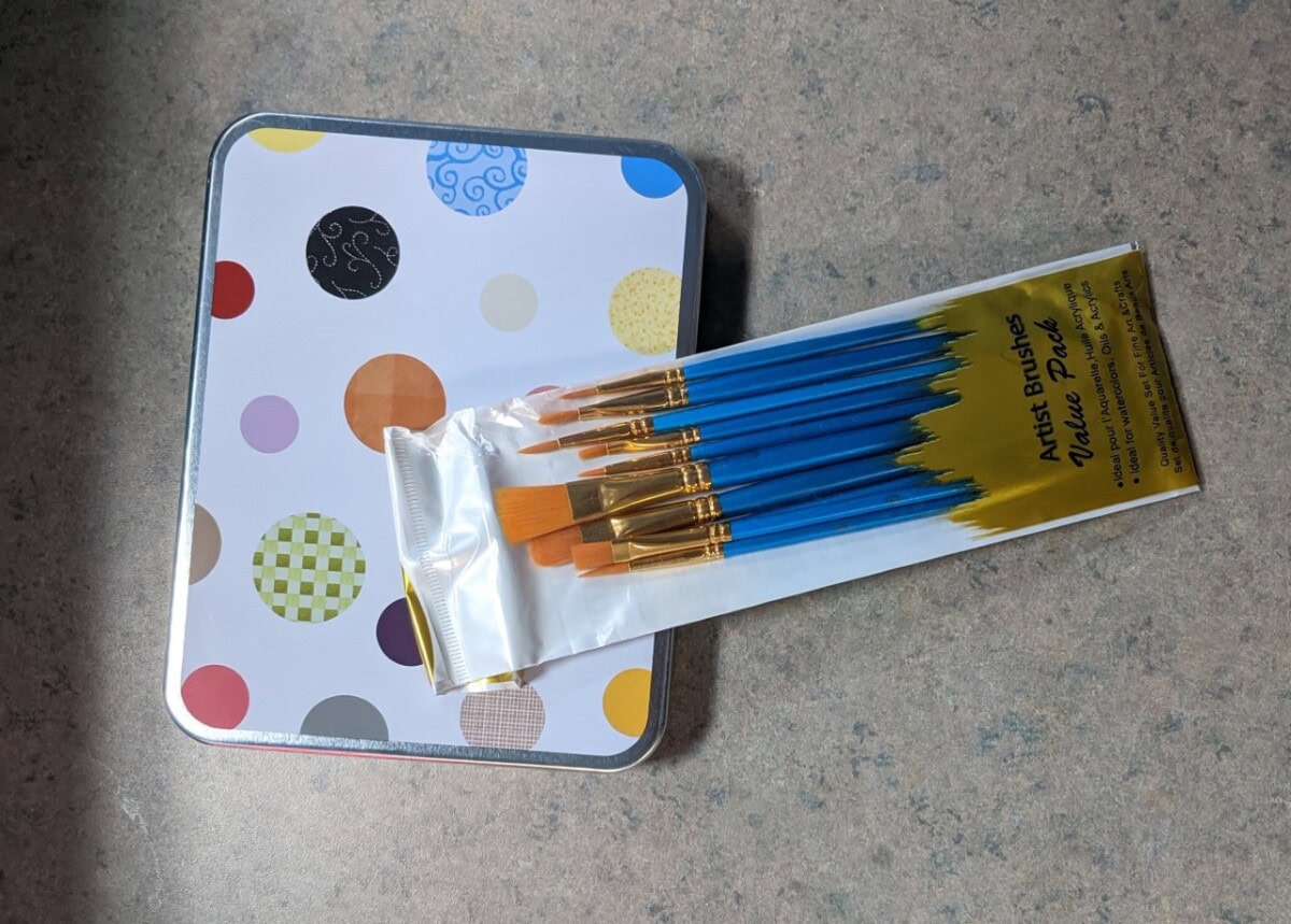 A set of blue-handled paintbrushes in plastic packaging rests on a polka-dotted tin box placed on a gray countertop.