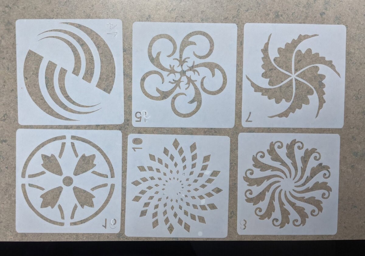 Six white square stencils with intricate cutout patterns, each featuring a unique abstract or floral design, arranged in two rows of three on a speckled beige surface. Numbers appear on some stencils.