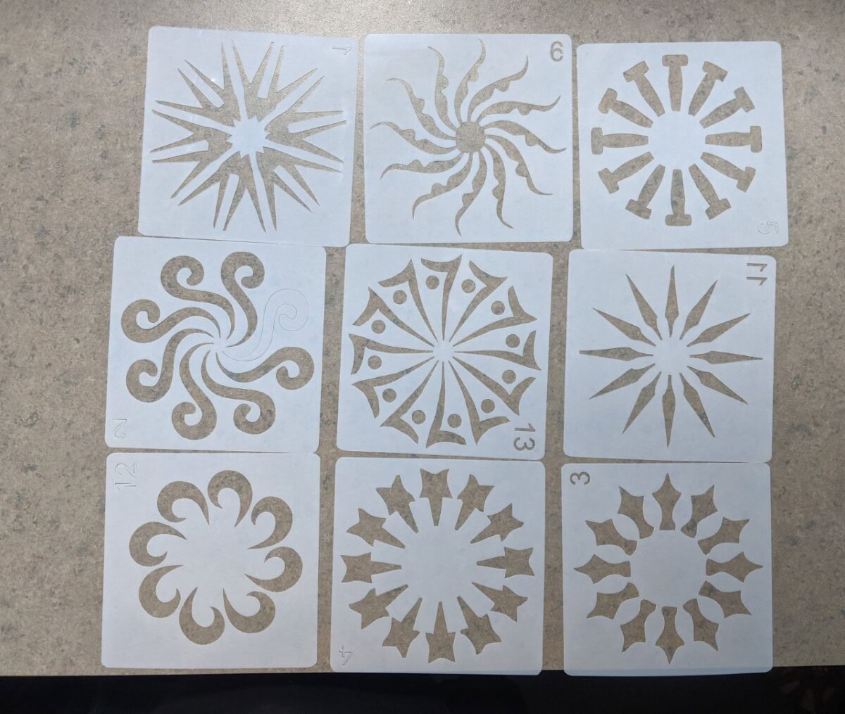 Nine square stencil cards with various symmetrical sunburst and swirl designs, each labeled with a number in the corner, are arranged in a 3x3 grid on a beige surface.