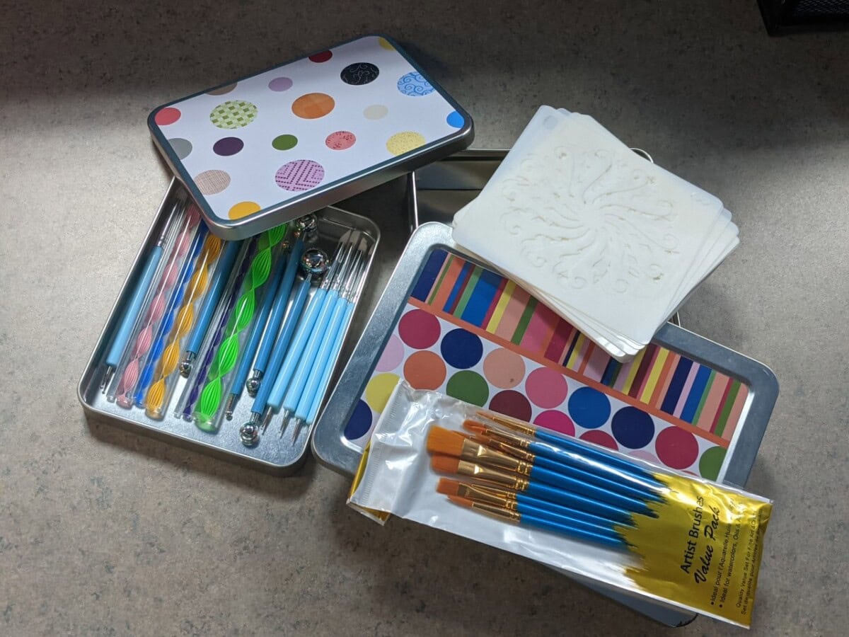 Art supplies on a table, including two decorative metal tins, dotting tools, paintbrushes in packaging, white coasters with embossed designs, and colorful striped and dotted cards.